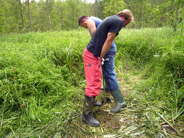 Two men in work clothes are leaning against a metal rod sticking out of the ground. Around them are trampled vegetation