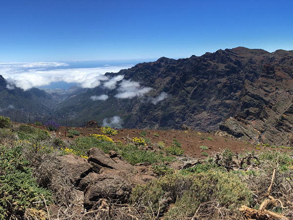 A view from high above of dark rocky mountains, a distant valley and a sea covered with white clouds. Blue sky above