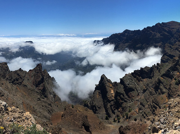 A view from high above the valley covered with white clouds. Dark, rocky peaks protrude above the clouds. Blue sky above