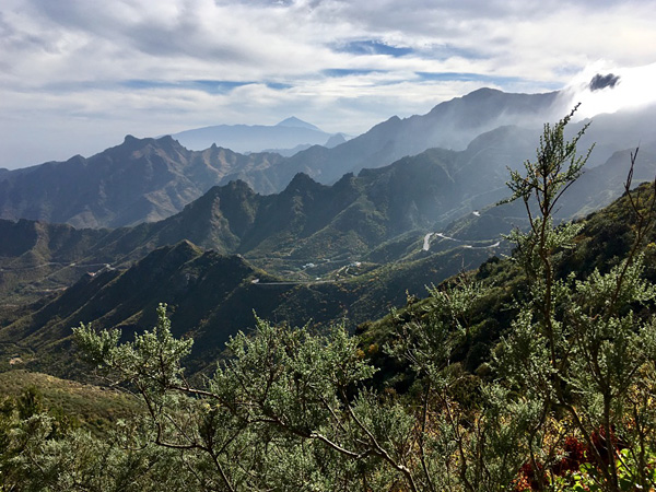 A view from high above of several mountain ranges lying one behind the other. In the foreground, sparse shrubby vegetation