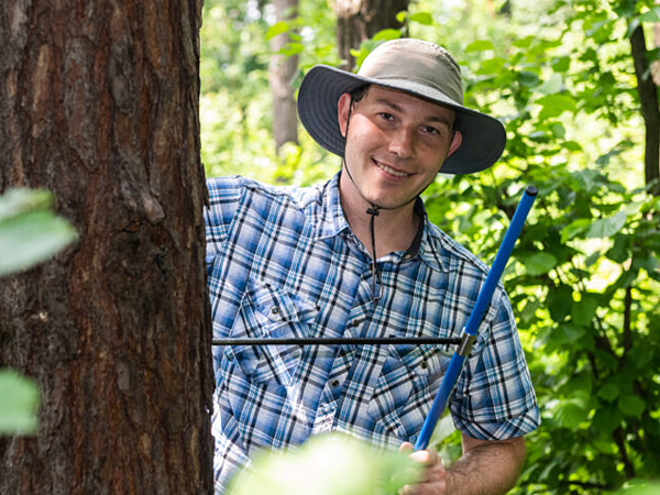 A man in a hat holds a metal auger stuck in a tree trunk by its handle