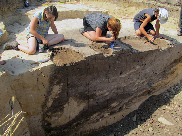 Three people kneel and use small shovels to scoop a thin layer of soil from the surface of the exposed soil profile