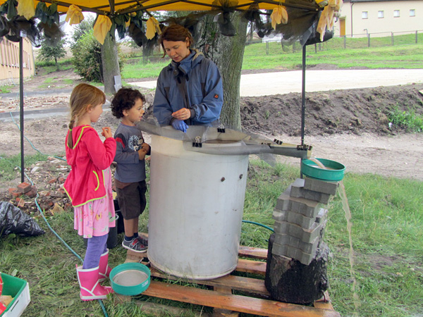 A woman stands behind a barrel-shaped device through which water flows. A boy and a girl watch her work