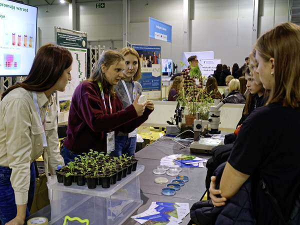 Three women show some students dishes with microorganism cultures. In front of them on the table are pots with small plants