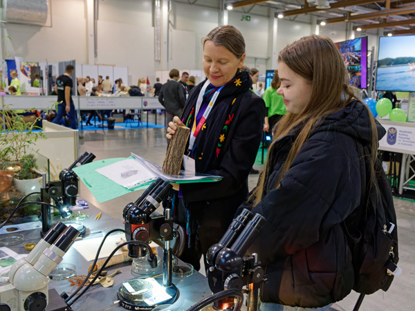 A woman stands at a table full of microscopes. She shows a student a piece of wood. In the distance, other exhibition stands