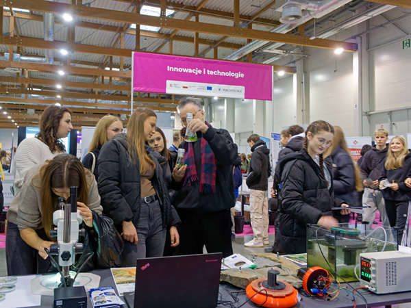 A man holds up a flask of green liquid, explaining something to several young women. In the background, a crowd of visitors